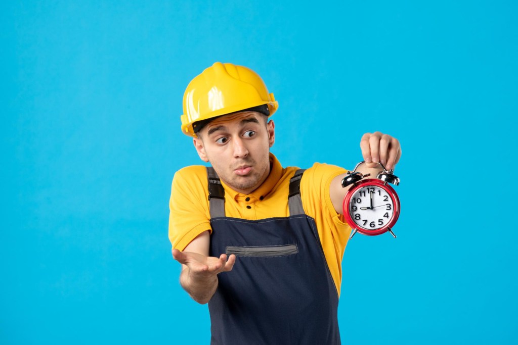 Ein Bauarbeiter mit einem gelben Helm hält eine Uhr in der Hand die 9 Uhr anzeigt. Er schaut verwundert auf die Uhr und macht dazu eine fragende Handbewegung da das Personalmanagement nicht funktioniert.