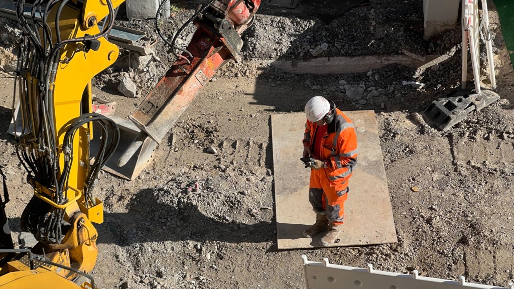Foto aus der Luft von einem Monteur in orange-farbener Baustellenkleidung mit weißem Schutzhelm. Er hat ein Smartphone in der Hand und dokumentiert gerade in der Vaira App. Links ragt außerdem ein gelber Baggerarm ins Bild.