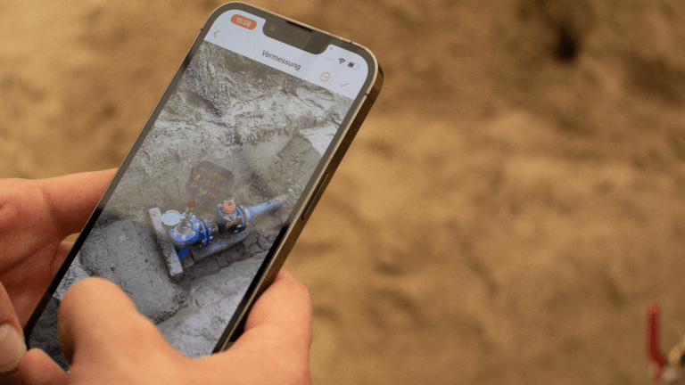 Zwei Hände ragen über einer Baustelle links ins Bild und halten ein Smartphone mit der Vaira App in der Hand. Mit der App wird eine Vermessung per Augmented Reality durchgeführt. Man erkennt einen abgelegten Messpunkt in AR sowieo einige Messwerte, die auf dem Display direkt angezeigt werden.