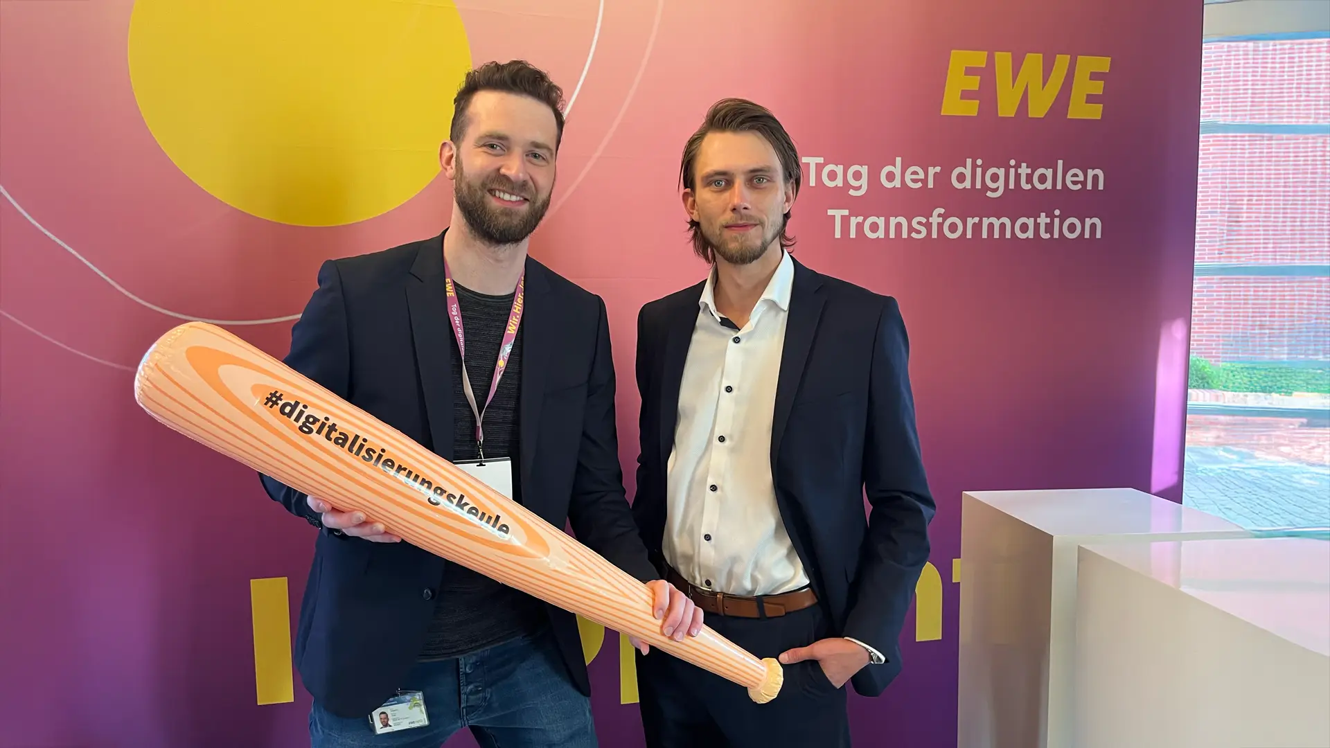 Uwe Kopplin (EWE Netz) und Philipp Buttmann (Vaira) posieren gemeinsam vor einem Event-Banner des EWE Digi Day 2024. Uwe hält eine Digitalisierungskeule von Vaira in der Hand.