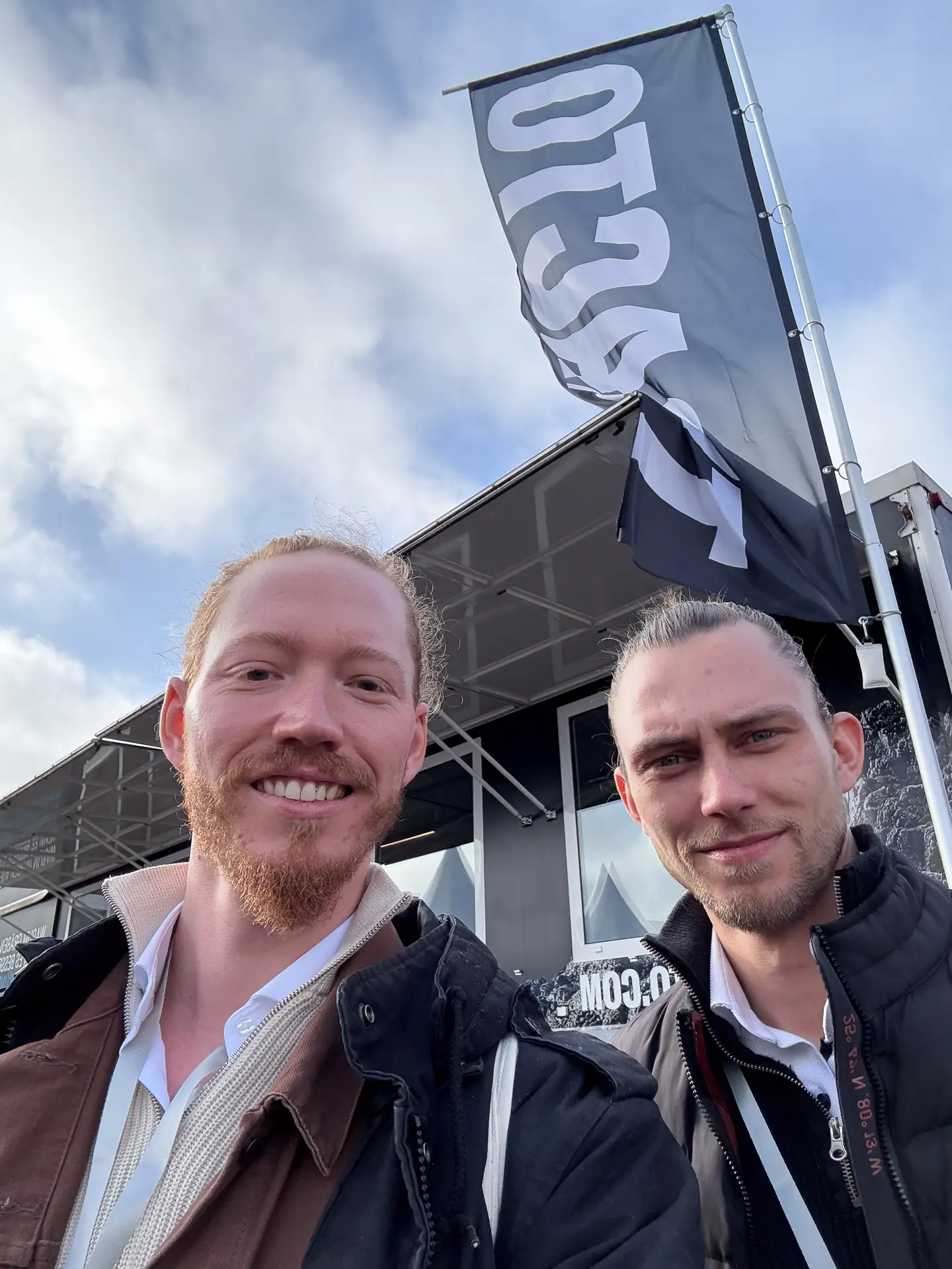 Selfie von Max und Philipp draußen auf der IRO 2025. Im Hintergrund weht eine Fahne von TRACTO.