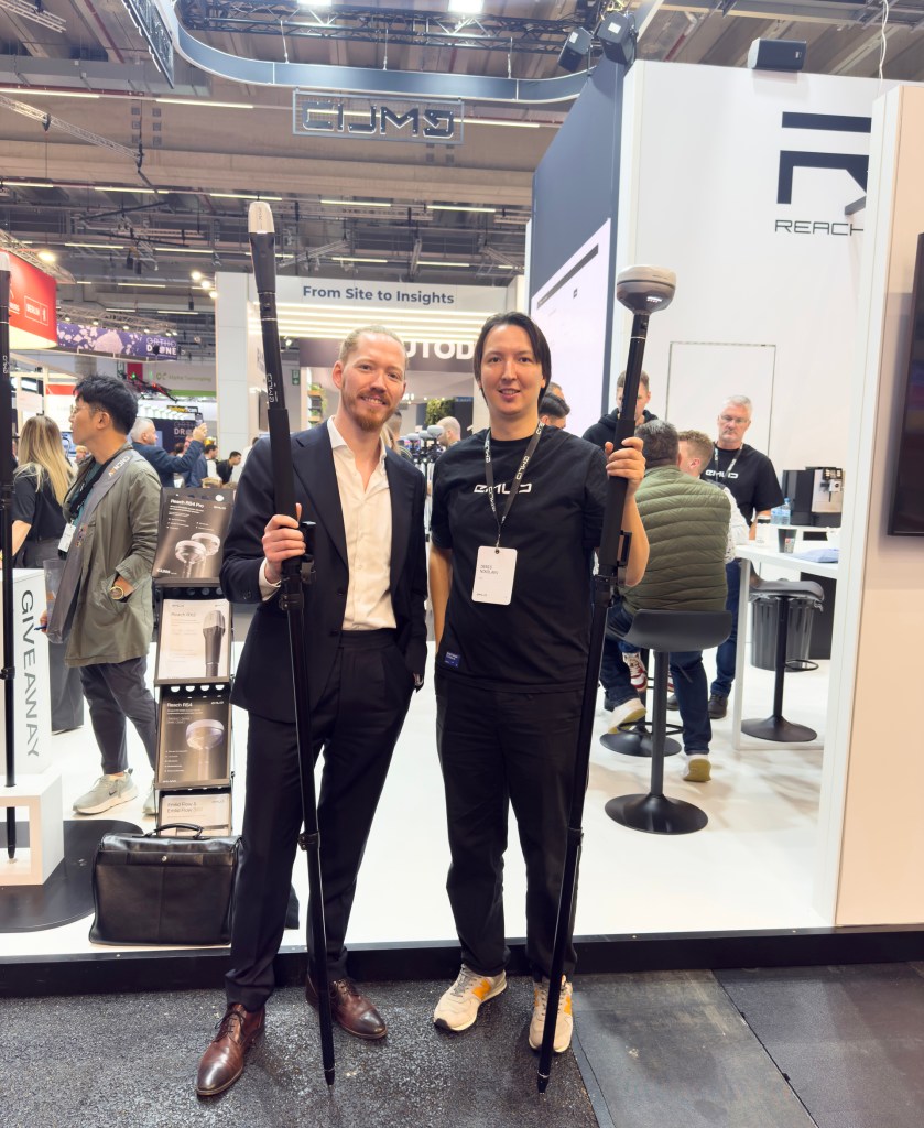 (v.l.) Max Erdmann (Vaira UG), Denis Nikolaev (Emlid). Beide halten die neusten Innovationen von Emlid in der Hand: Max hält eine Emlid Reach RX 2 in der Hand, Denis eine Reach RS4. Beide sind an langen Stäben befestigt.